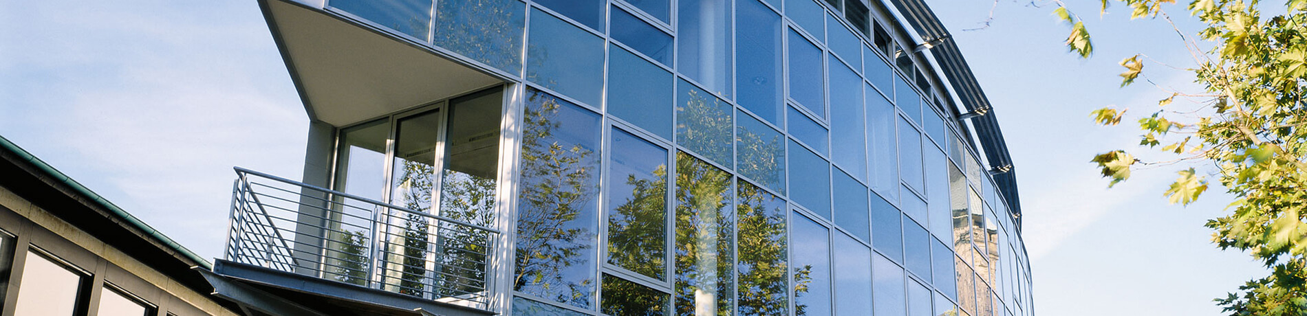 Nahaufnahme der geschwungenen Glasfront eines modernen Bürogebäudes mit Balkon und Bäumen.