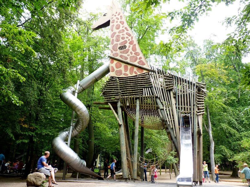 Große hölzerne Kletterstruktur in Form einer Giraffe auf einem Waldspielplatz mit langer Röhrenrutsche.