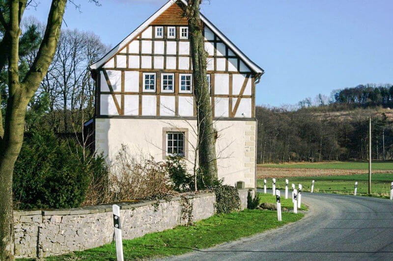 Historisches Fachwerkhaus an einer Straßenkurve mit Natursteinmauer, Leitpfosten und ländlicher Umgebung bei Sonnenschein.