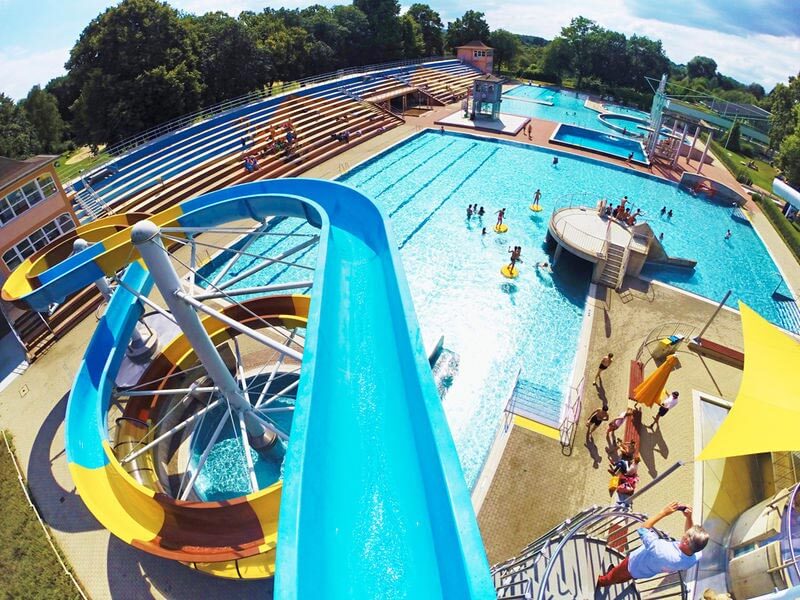 Belebtes Freibad in Vogelperspektive mit blauer Wasserrutsche, Schwimmbecken und Tribüne an einem Sonnentag.