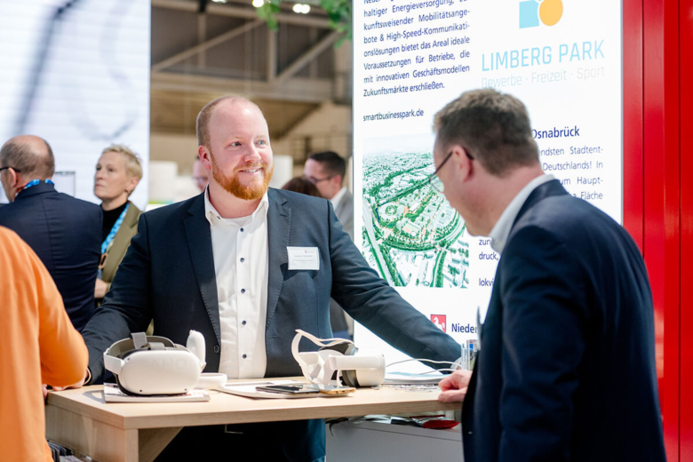 Ein Mann im Anzug im Gespräch an einem Messestand der Wirtschaftsförderung mit VR-Brillen auf dem Tisch.
