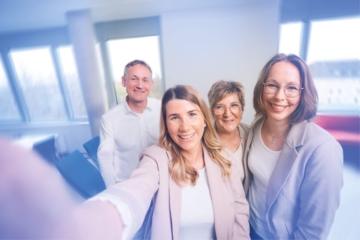Vier lächelnde Personen aus einem Büroteam machen gemeinsam ein Nahaufnahme-Selfie in einem Besprechungsraum.