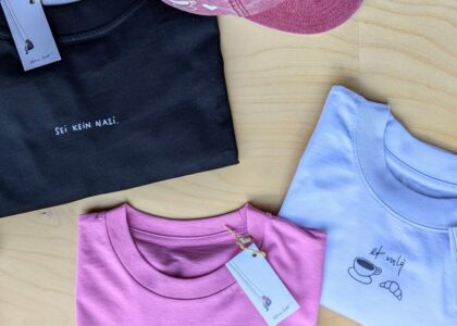 Gefaltete T-Shirts in Schwarz, Rosa und Weiß mit Sprüchen sowie eine rosa Basecap auf einem Holzhintergrund.