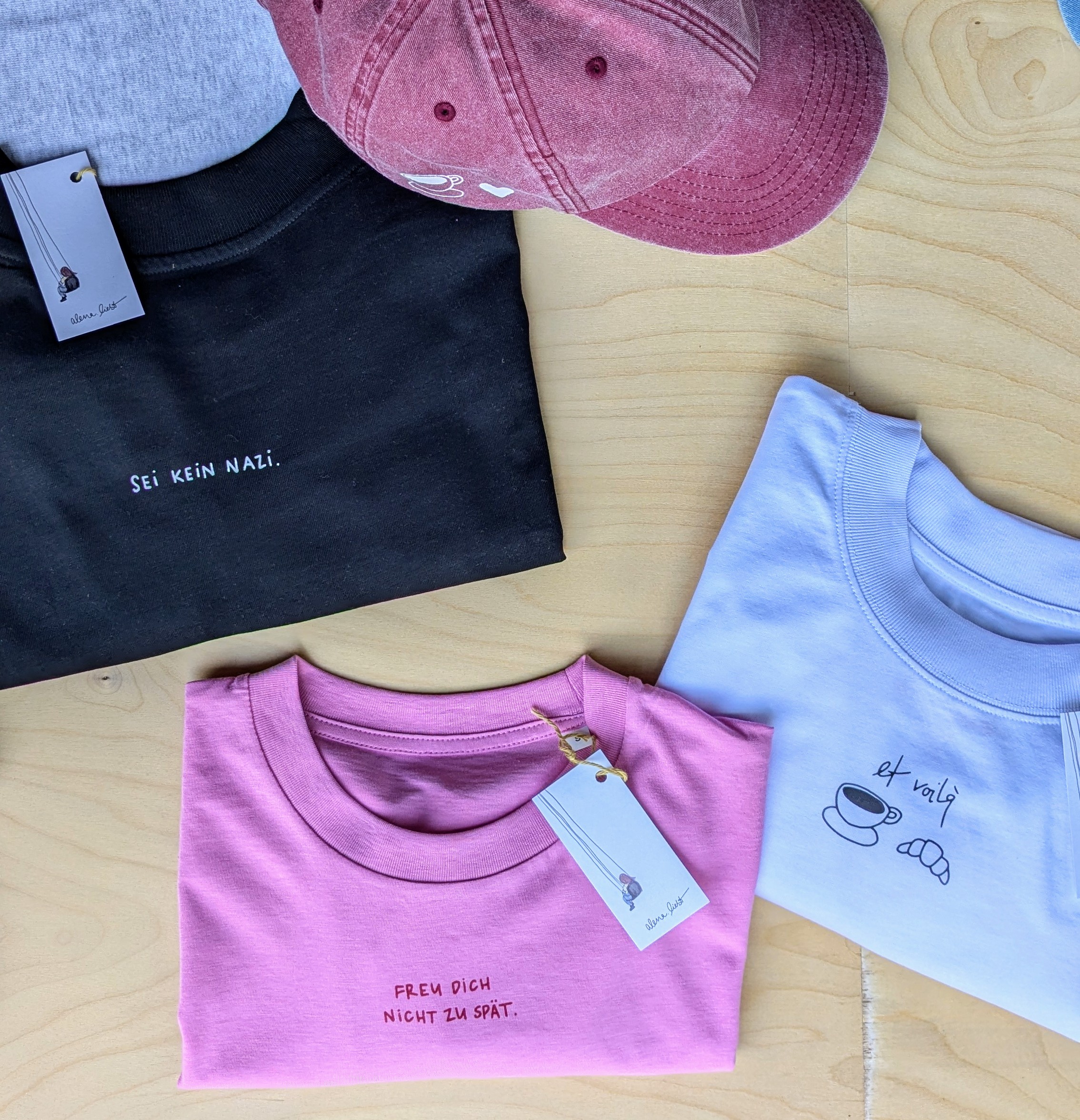 Gefaltete T-Shirts in Schwarz, Rosa und Weiß mit Sprüchen sowie eine rosa Basecap auf einem Holzhintergrund.