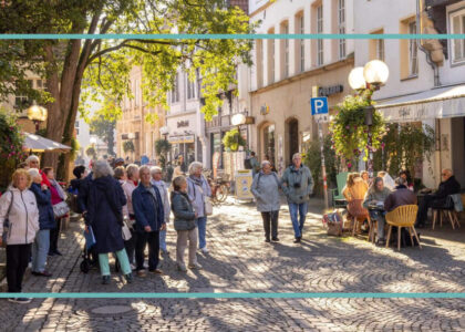 Zahlreiche Menschen gehen bei Sonnenschein über eine alte Kopfsteinpflasterstraße mit schattigen Bäumen und historischen Fachwerkhäusern.