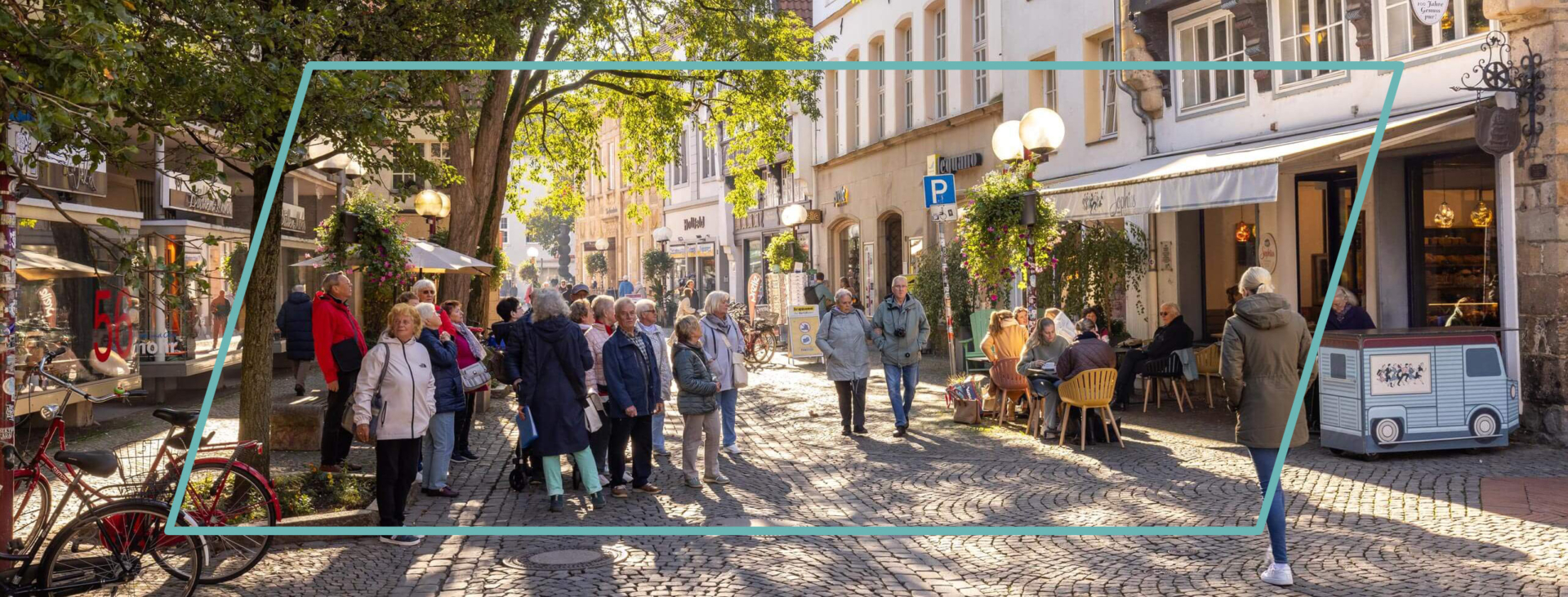 Zahlreiche Menschen gehen bei Sonnenschein über eine alte Kopfsteinpflasterstraße mit schattigen Bäumen und historischen Fachwerkhäusern.