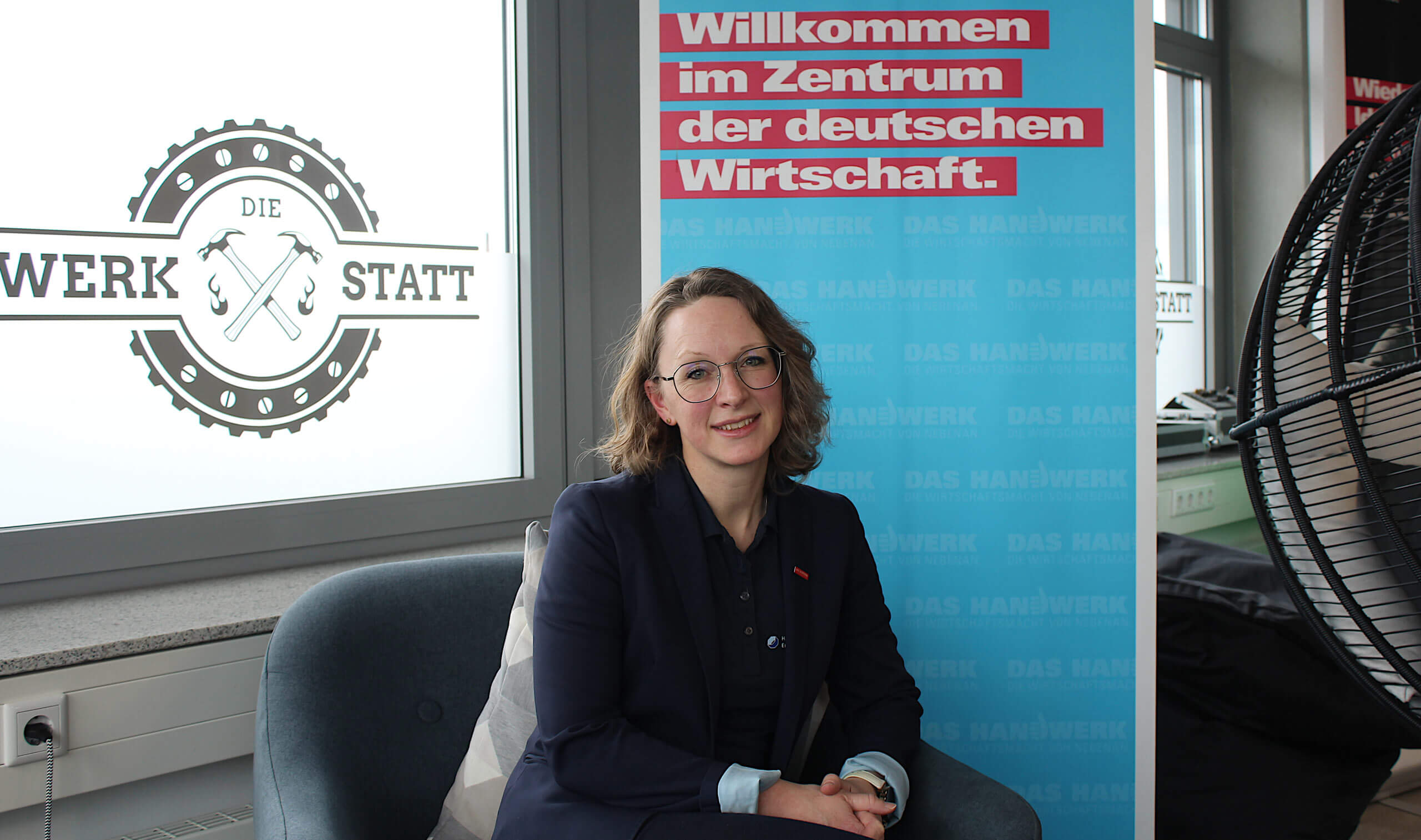 Lächelnde Frau mit Brille im Blazer sitzt in einem Sessel vor einem Plakat mit der Aufschrift „Willkommen im Zentrum der deutschen Wirtschaft“.