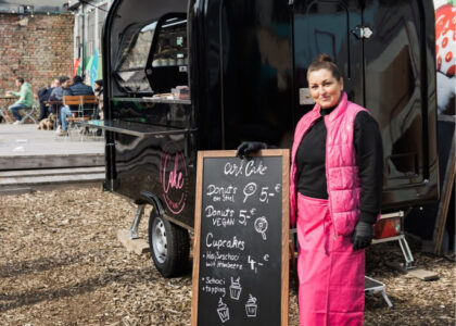 Eine Frau in pinkfarbener Veste steht lächelnd neben einer schwarzen Kreidetafel vor einem schwarzen Foodtruck für Gebäck.