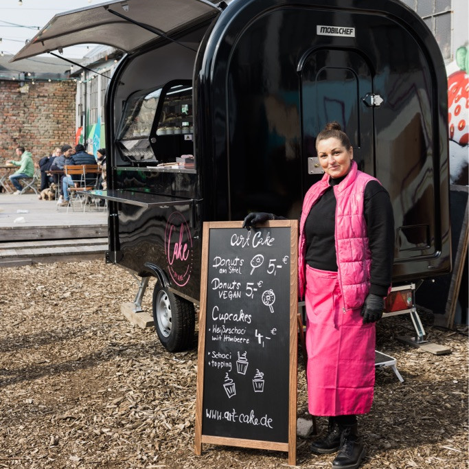 Eine Frau in pinkfarbener Veste steht lächelnd neben einer schwarzen Kreidetafel vor einem schwarzen Foodtruck für Gebäck.