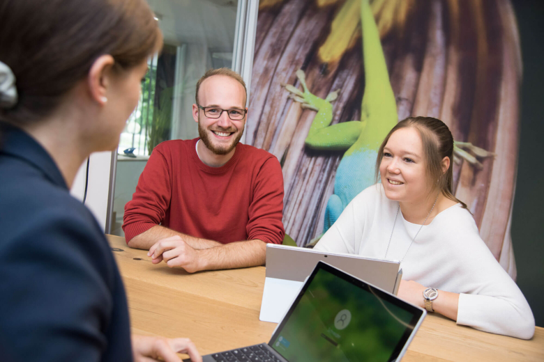 Zwei junge Erwachsene sitzen lächelnd an einem Tisch mit Laptops und sprechen mit einer Beraterin.