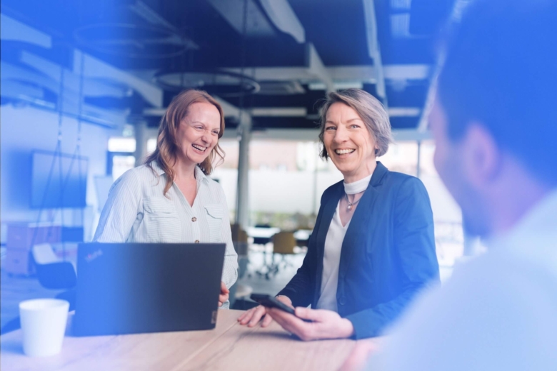 Zwei lächelnde Frauen im Business-Outfit unterhalten sich in einem modernen Büro an einem Tisch mit Laptop.