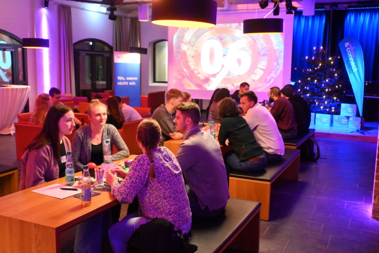 Junge Menschen sitzen in Gruppen an Holztischen und beratschlagen sich während eines Quiz-Events in einem Raum.