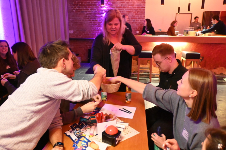 Eine Frau lässt Personen an einem Tisch Loszettel aus einer Schale ziehen, auf dem Tisch liegen Quiz-Utensilien.