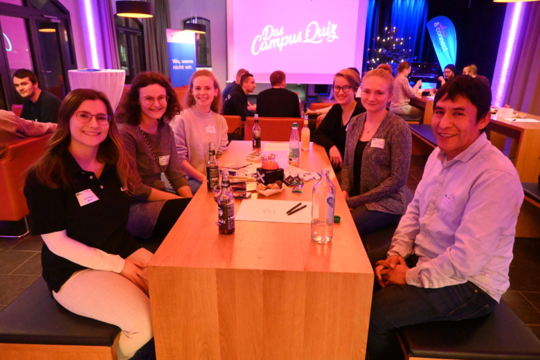 Eine Gruppe von sechs Personen sitzt gemeinsam an einem Holztisch bei einem Quiz-Event und lächelt in die Kamera.