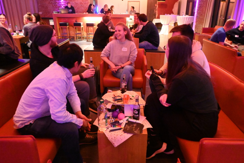 Eine Gruppe junger Menschen sitzt in einer Lounge-Ecke zusammen, unterhält sich und hat Getränke auf dem Tisch.