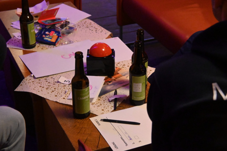 Tisch mit Bierflaschen, Lösungszettel, Stift und einem roten Quiz-Buzzer bei einer Event-Veranstaltung.
