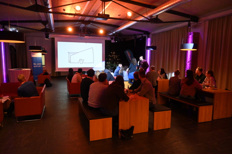 Blick in den Veranstaltungsraum, in dem Publikum auf Bänken sitzt und auf die Quiz-Leinwand schaut.