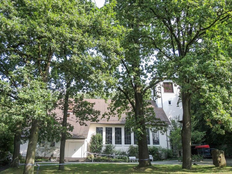 Weißes Kirchengebäude mit rotem Ziegeldach, umgeben von hohen, dicht bewachsenen Laubbäumen.