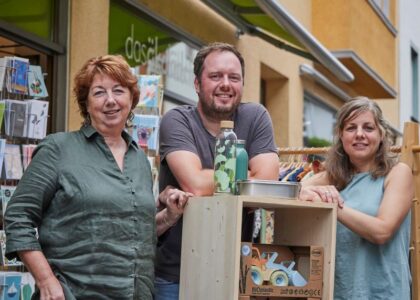Drei lächelnde Personen stehen vor einem Bioladen neben einem Regal mit nachhaltigen Produkten wie Biokunststoff-Spielzeug und Trinkflaschen.