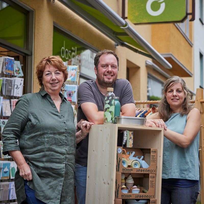 Drei lächelnde Personen stehen vor einem Bioladen neben einem Regal mit nachhaltigen Produkten wie Biokunststoff-Spielzeug und Trinkflaschen.