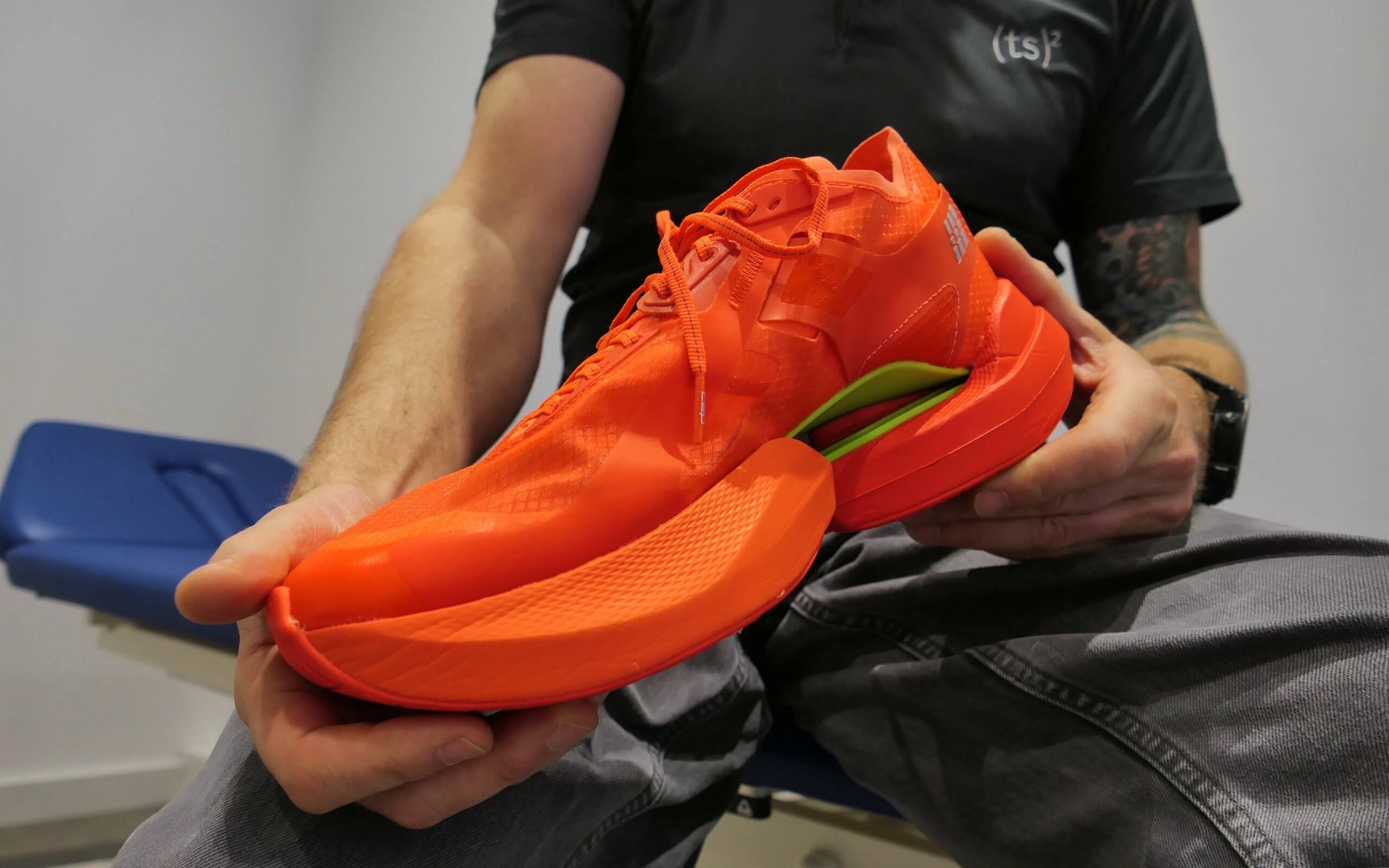 Hände halten einen auffällig leuchtend orangefarbenen Laufschuh nah vor die Kamera.