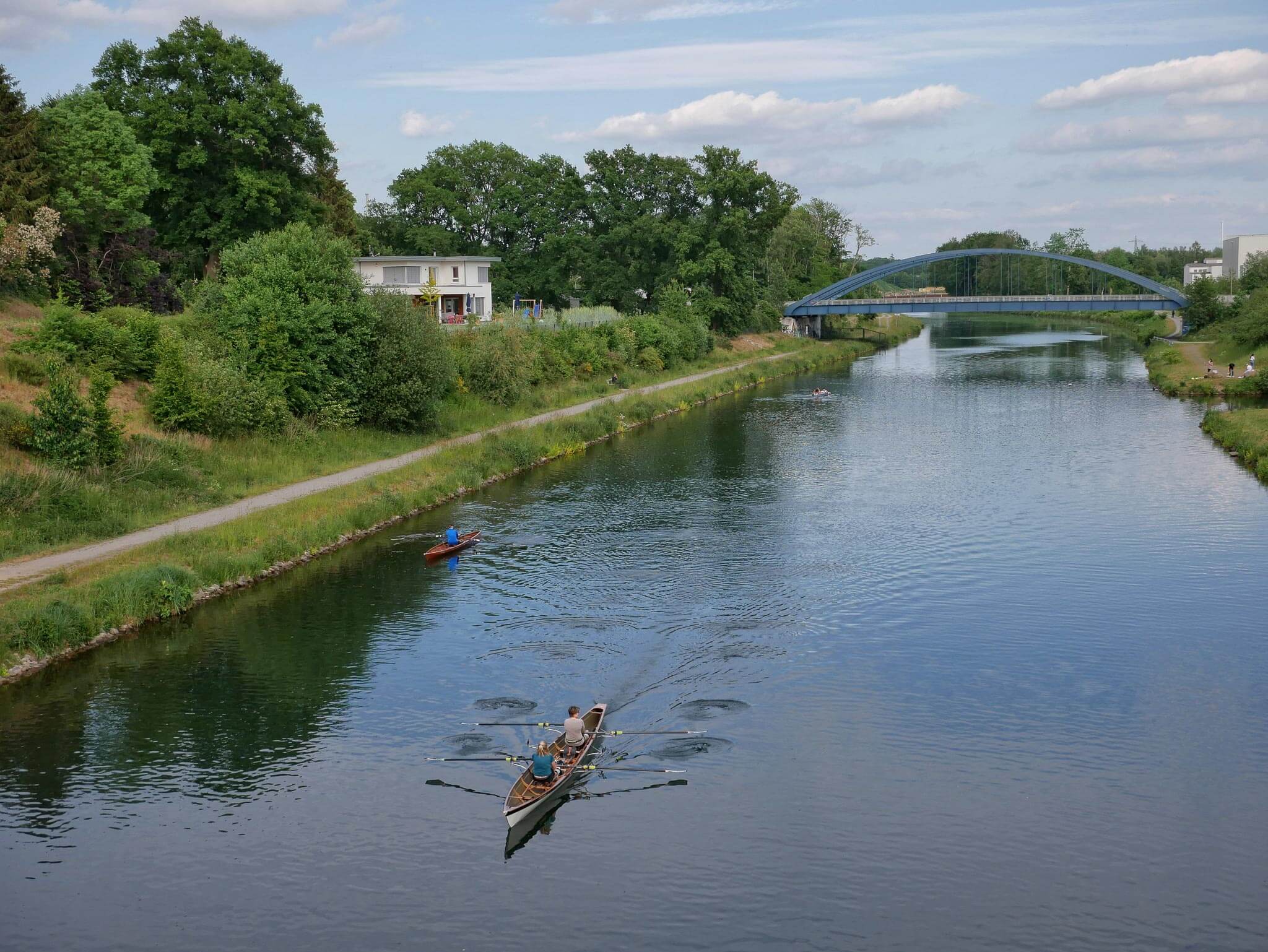 Ruderboot auf einem ruhigen Kanal mit grünen Ufern und einer blauen Bogenbrücke im Hintergrund.