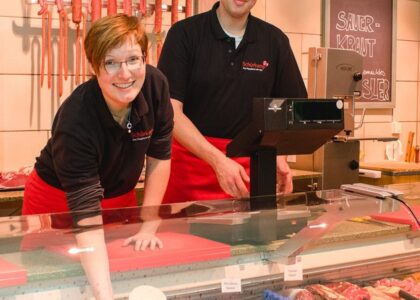 Zwei Metzger in „Schürkamp“-Uniform lächeln hinter einer gut gefüllten Wursttheke. Im Hintergrund hängen Würste.