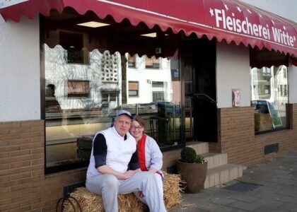Ein Paar sitzt lächelnd auf einem Strohballen vor dem Geschäftseingang der „Fleischerei Witte“ unter einer roten Markise.