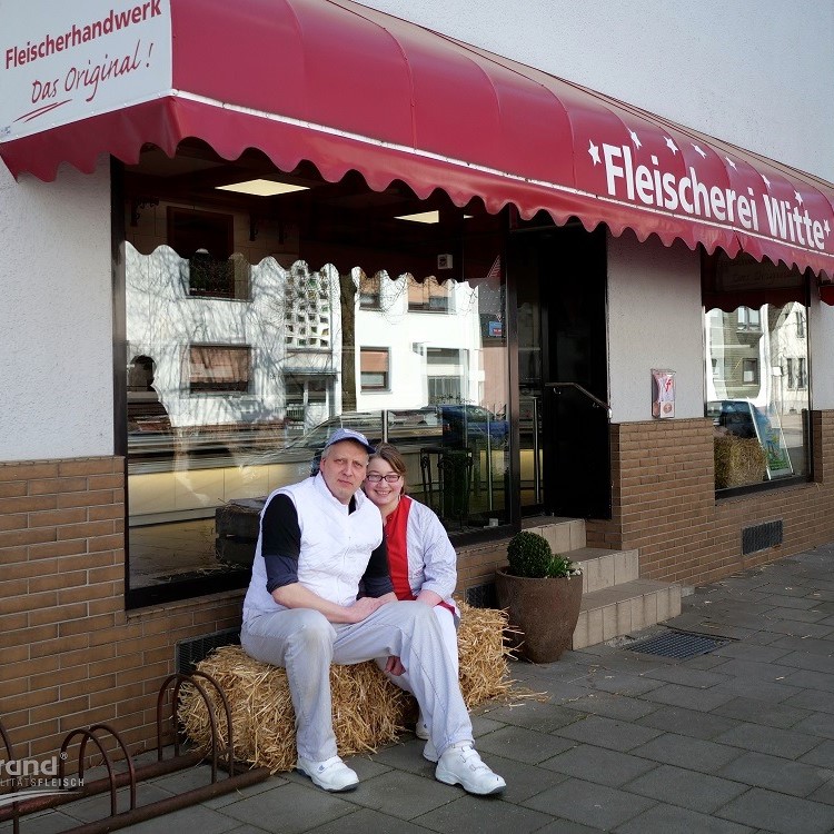 Ein Paar sitzt lächelnd auf einem Strohballen vor dem Geschäftseingang der „Fleischerei Witte“ unter einer roten Markise.