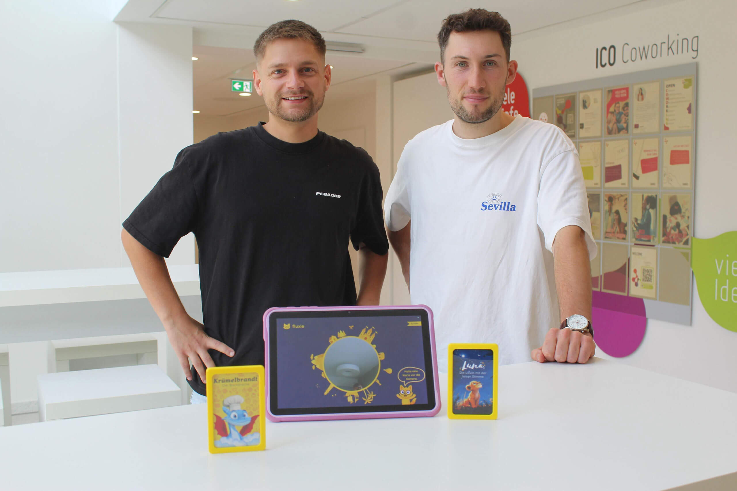 Zwei Männer stehen lächelnd hinter einem Tisch mit einem Tablet und gelben Lernkarten.