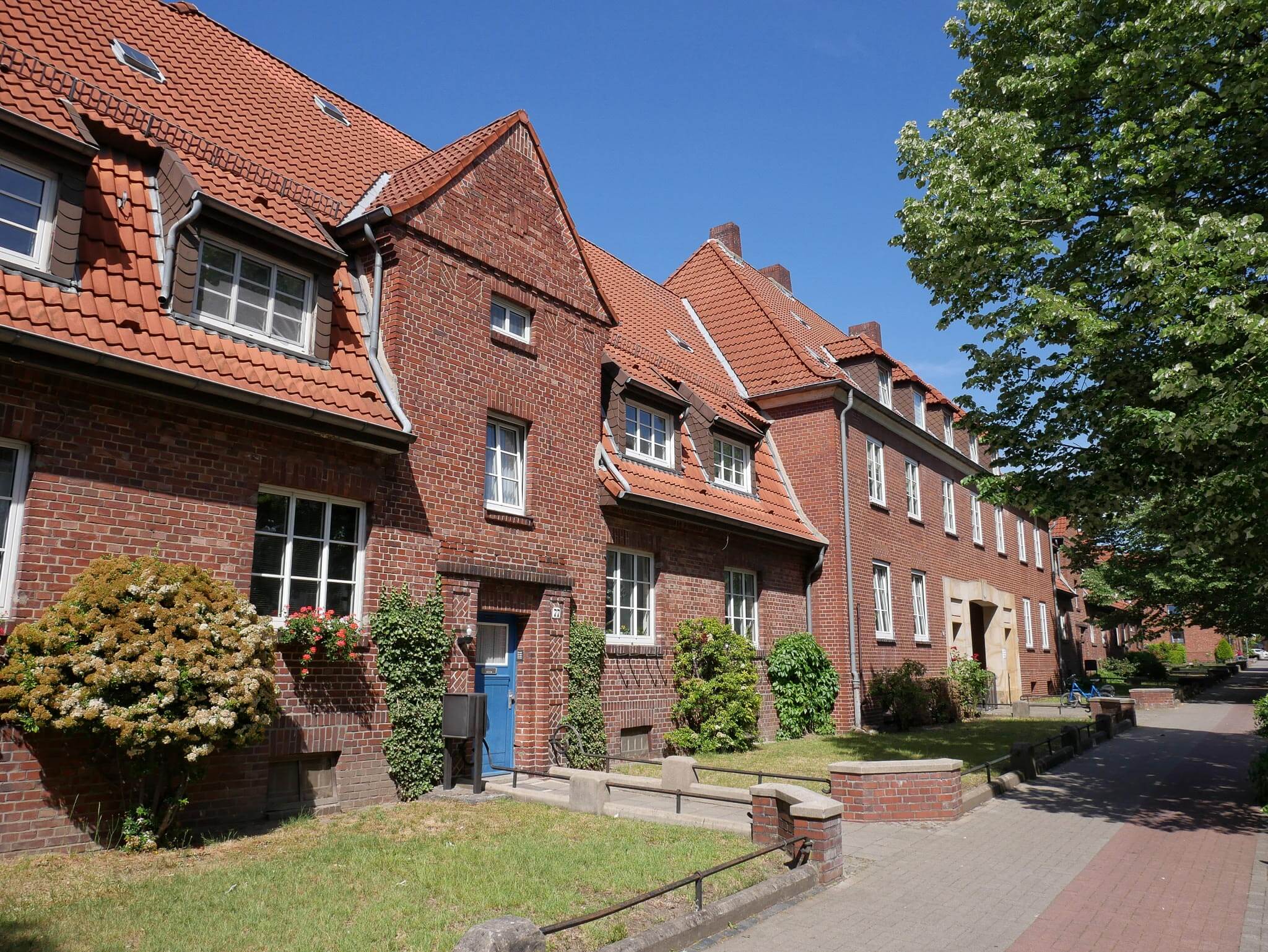 Mehrstöckige Backstein-Wohngebäude mit rotem Ziegeldach, kleinen Vorgärten und einem gepflasterten Gehweg.