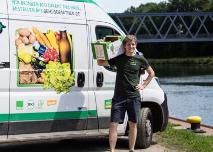 Ein Lieferant steht mit einer Kiste Bio-Gemüse lächelnd neben einem weißen Lieferwagen der Gemüsegärtner an einem Flussufer.