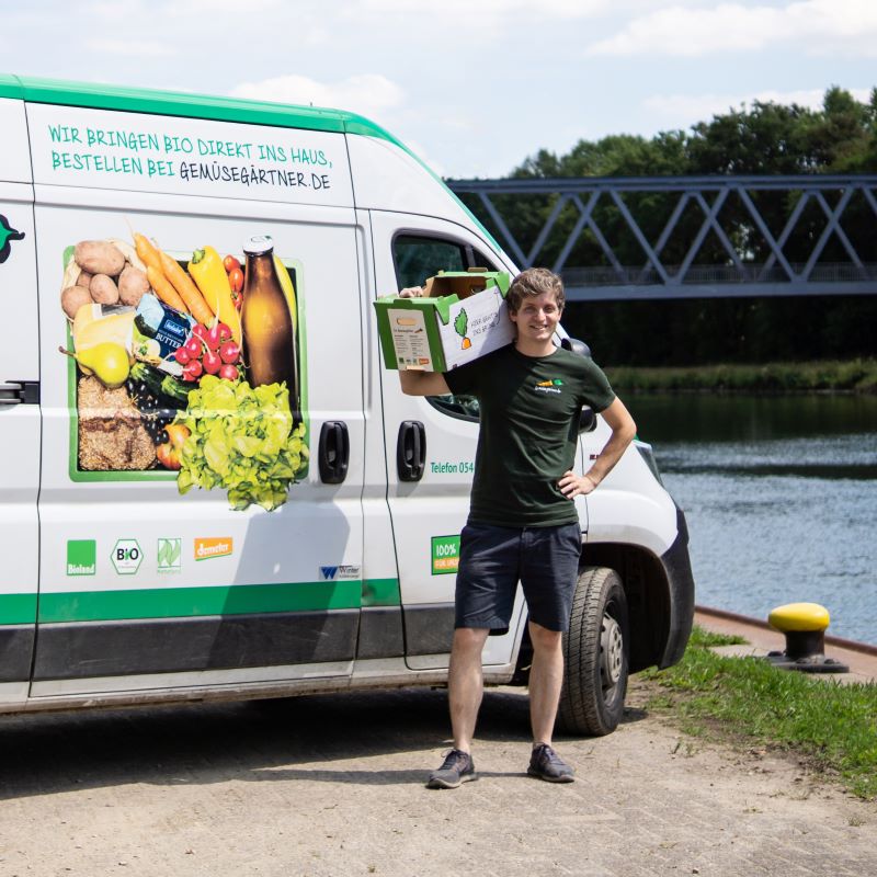 Ein Lieferant steht mit einer Kiste Bio-Gemüse lächelnd neben einem weißen Lieferwagen der Gemüsegärtner an einem Flussufer.