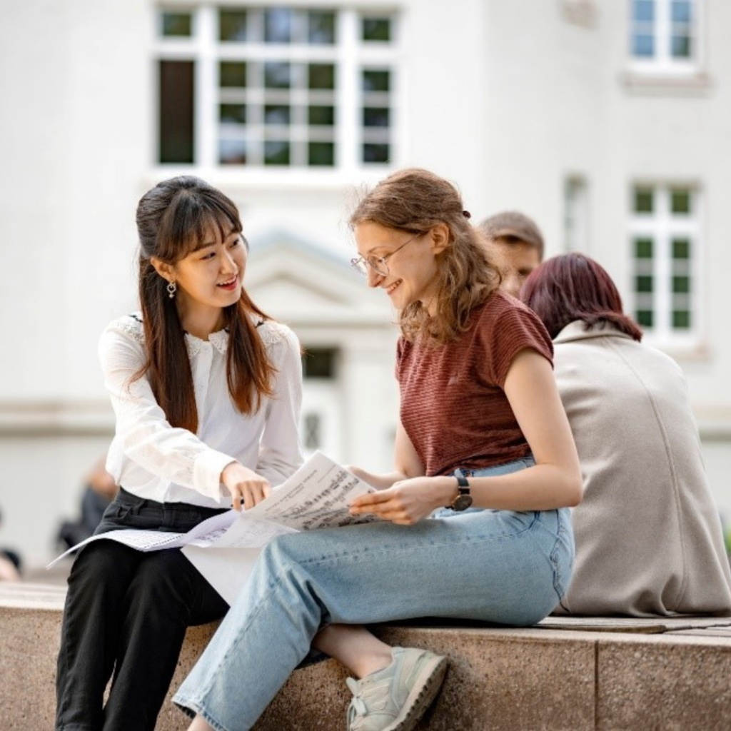 Zwei Studentinnen sitzen im Freien auf einer Steinmauer und betrachten gemeinsam Unterlagen auf einem Papierblatt.