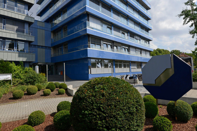 Mehrstöckiges blaues Bürogebäude der Handwerkskammer bei Sonnenschein mit zugeschnittenen Büschen im Vorgarten.