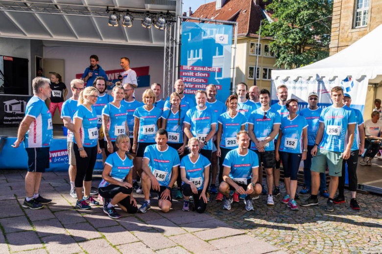 Eine große Laufgruppe in hellblauen Trikots mit Startnummern posiert vor einer Veranstaltungsbühne.