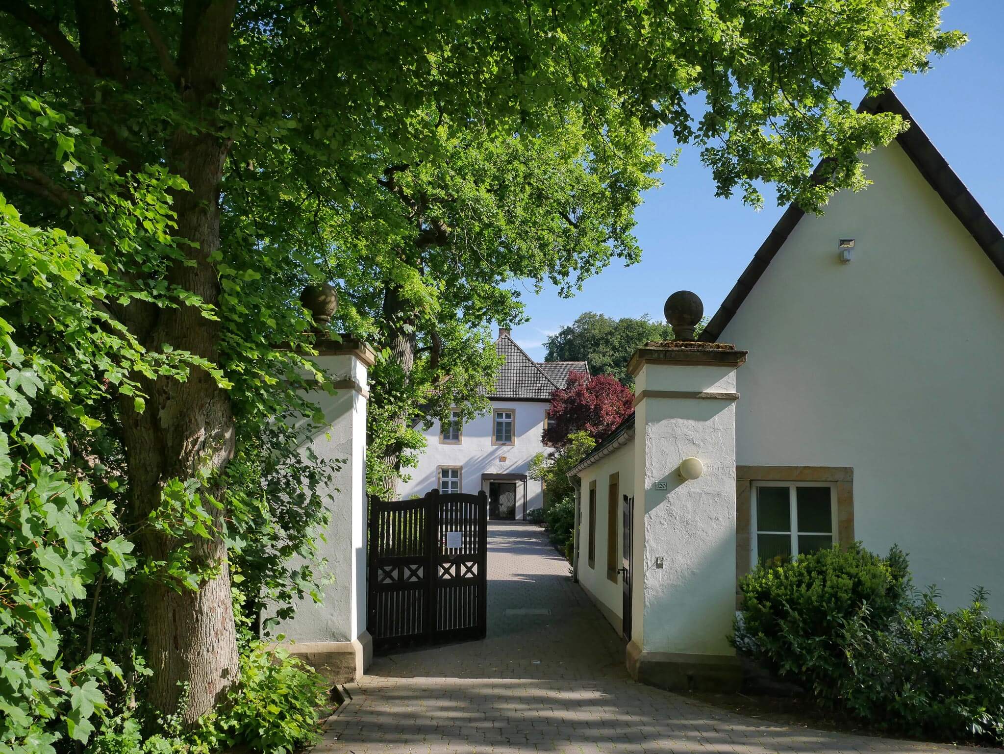 Offenes, dunkles Holztor zwischen weißen Torpfeilern, das den Blick auf ein historisches Gebäude freigibt.
