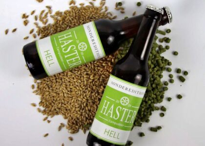Zwei Flaschen „Haster Campusbier Hell“ liegen auf Gerstenkörnern und Hopfenpellets auf weißem Hintergrund.