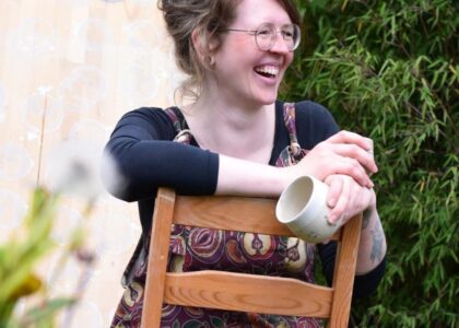 Eine Frau mit Brille und gemusterter Latzhose sitzt lachend auf einem umgedrehten Holzstuhl im Garten, eine Tasse haltend.
