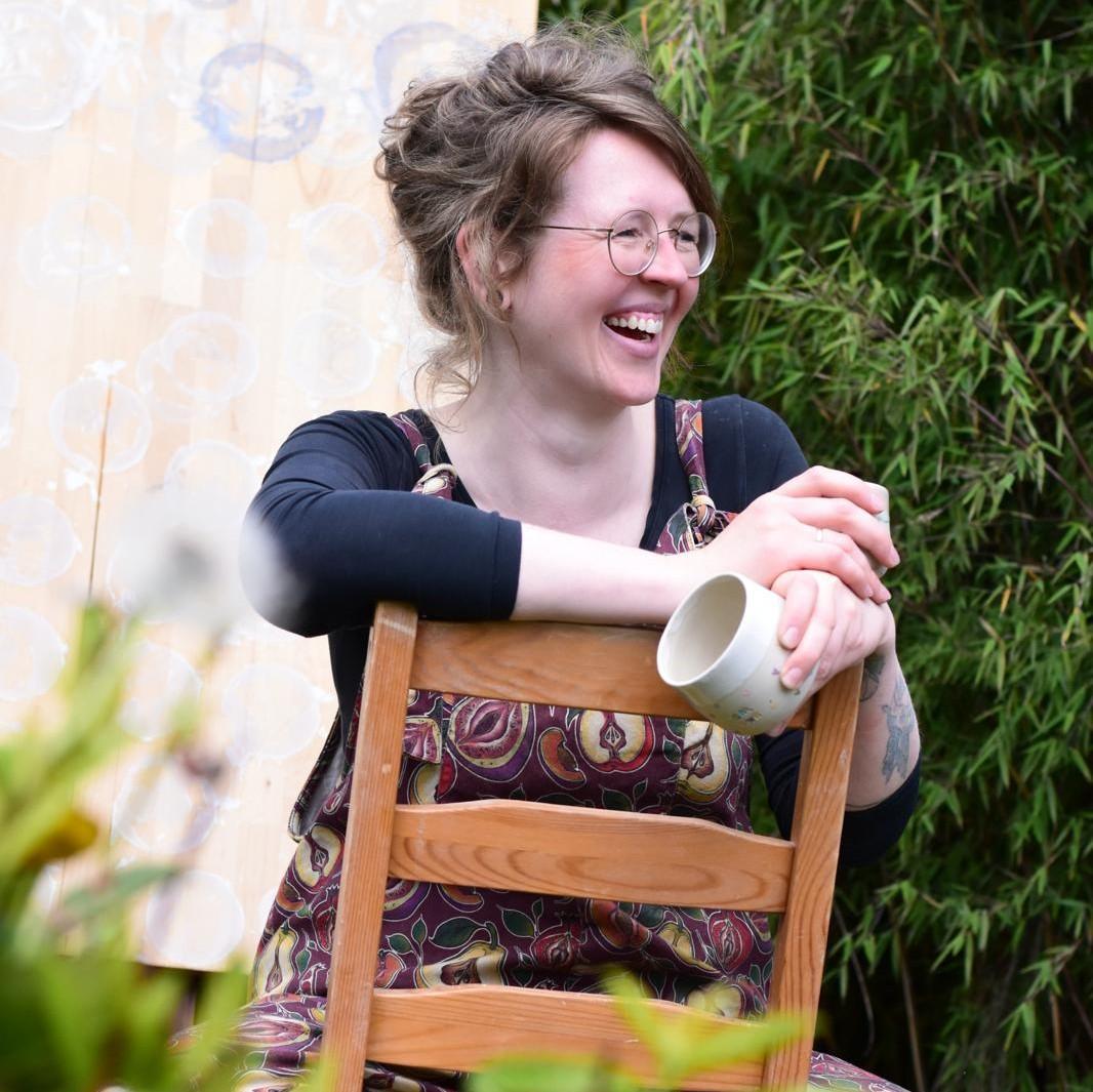 Eine Frau mit Brille und gemusterter Latzhose sitzt lachend auf einem umgedrehten Holzstuhl im Garten, eine Tasse haltend.