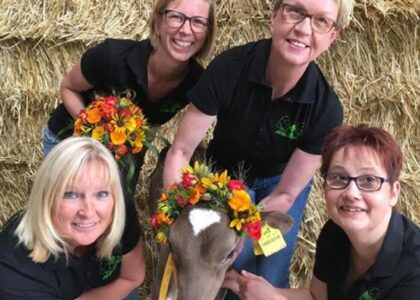 Vier lächelnde Frauen in schwarzen Poloshirts posieren mit einem geschmückten Kalb vor Strohballen.
