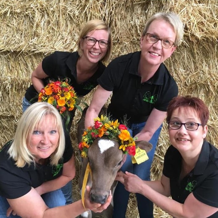 Vier lächelnde Frauen in schwarzen Poloshirts posieren mit einem geschmückten Kalb vor Strohballen.