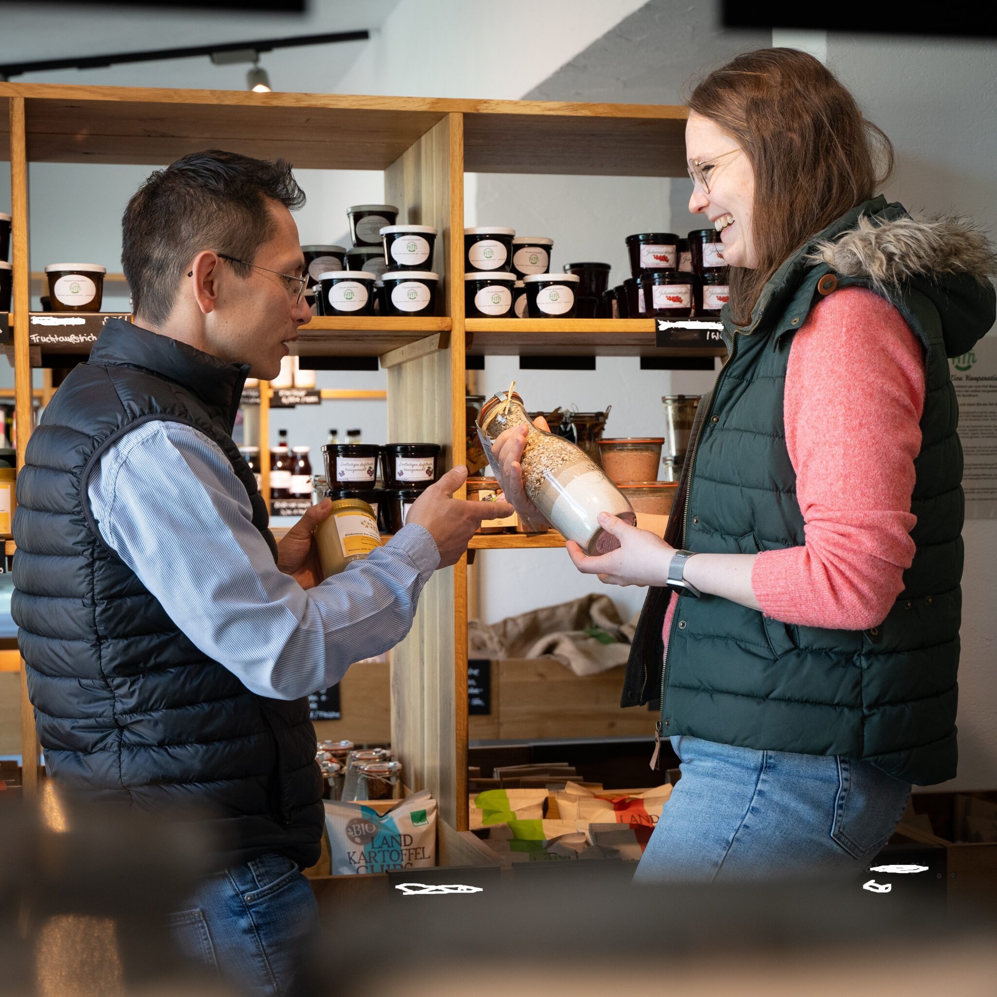 Ein Mann und eine Frau unterhalten sich lächelnd in einem Hofladen und betrachten Produkte in Gläsern vor vollen Regalen.