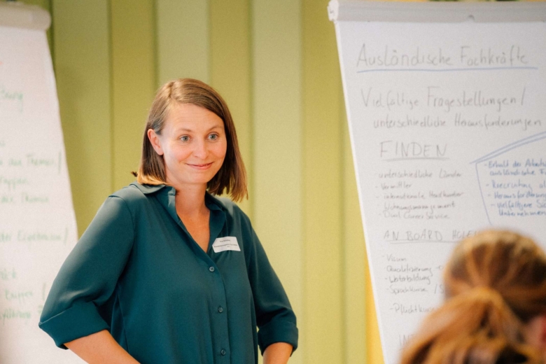 Lächelnde Frau im grünen Hemd steht neben einem Beschrifteten Flipchart im Seminarraum.