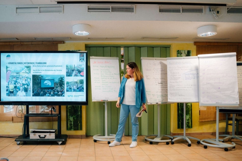Frau im blauen Hosenanzug präsentiert vor einem großen Monitor und mehreren Flipcharts.
