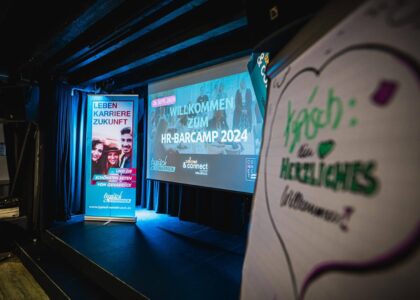 Veranstaltungsbühne mit Roll-up-Banner und Leinwandprojektion des „HR-Barcamp 2024“ vor blauer Beleuchtung.