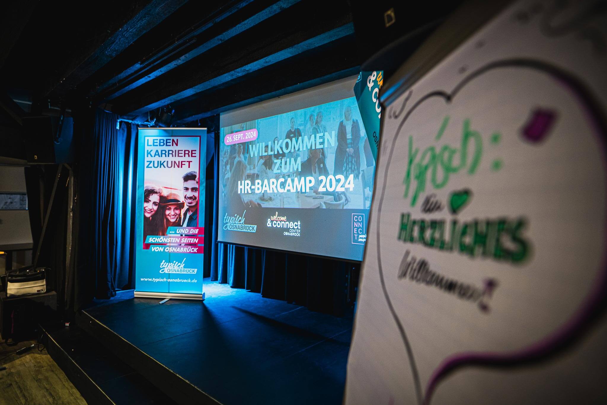 Veranstaltungsbühne mit Roll-up-Banner und Leinwandprojektion des „HR-Barcamp 2024“ vor blauer Beleuchtung.