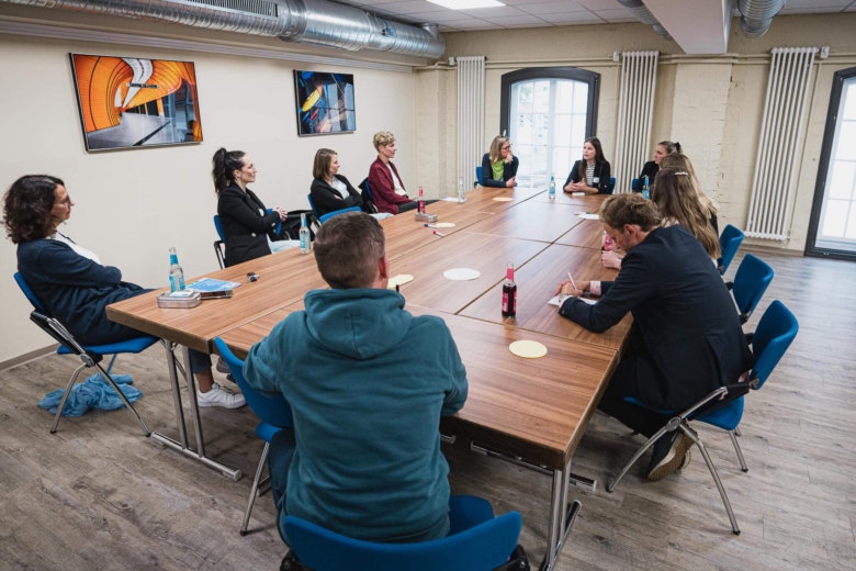 Menschengruppe sitzt in U-Form an einem großen Holztisch zusammen und führt eine Diskussion.