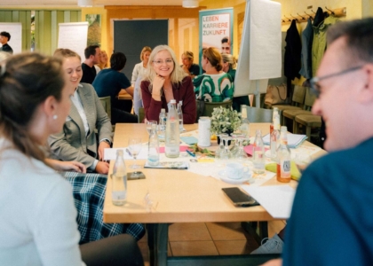 Lächelnde Menschen sitzen während eines Workshops im Gespräch an einem gedeckten Holztisch.