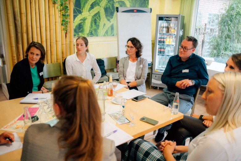 Teilnehmende einer Konferenz sitzen im Kreis um einen Tisch und hören aufmerksam zu.